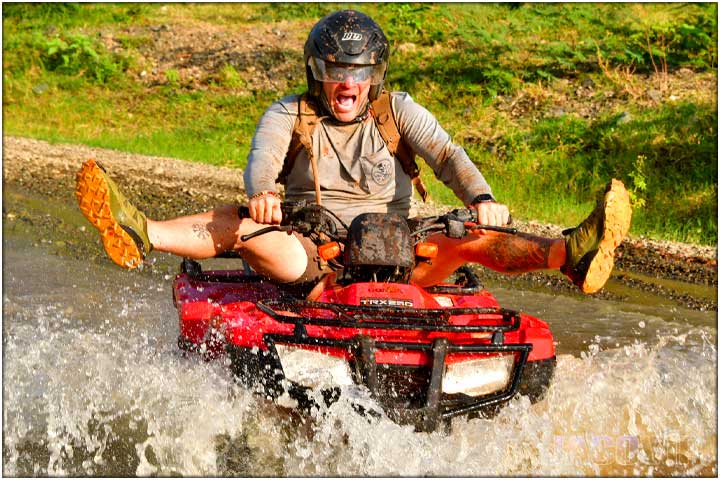 Guy on ATV splashing across river with legs up in the air